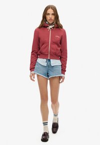 Sudadera roja con cremallera y cordón blanco, superpuesta a una camiseta azul clara, combinada con pantalones cortos de mezclilla y mocasines oscuros.