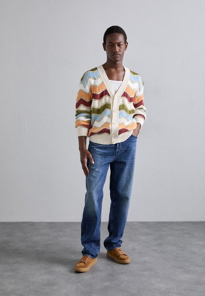 Adīts kardigans ar zigzaga rakstu krēmkrāsā, oranžā, zaļā un zilā krāsā, kombinēts ar zilajiem džinsiem un pelēkajiem sporta apaviem.