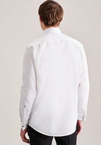 Camicia bianca a maniche lunghe con texture liscia, colletto classico, orlo arrotondato e polsini con bottoni, arricchiti da dettagli decorativi.