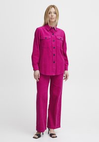 Fuchsia Corduroy-Hemd mit zwei Brusttaschen und schwarzen Knöpfen, kombiniert mit passenden weit geschnittenen Hosen. Schwarze offene High Heels.