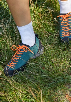 Teal og svart semsket skot med oransje snøring, med en teksturert såle, iført hvite ribbestrikkede sokker, på gressete underlag.