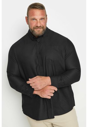 Mann mit Bart trägt ein schwarzes Hemd mit Knöpfen und eine helle Hose, steht vor einem schlichten weißen Hintergrund und blickt mit einem leichten Lächeln nach vorn.