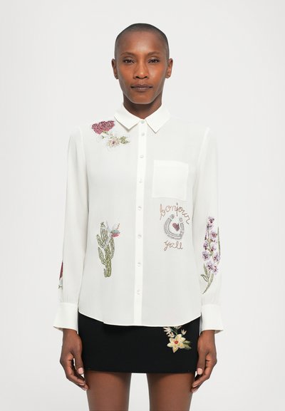 Weiße Bluse mit floralem Stickmuster in Rot, Grün und Lila. Knopfverschluss, Fronttasche, kombiniert mit einem schwarzen Rock mit floralen Akzenten.