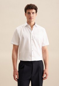 Camicia bianca a maniche corte con colletto button-down e taschino sul petto. Presenta una texture liscia e un tessuto leggero, indossata con pantaloni scuri.