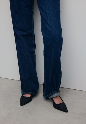 Pointed black woven heels paired with dark blue flared jeans, featuring visible stitching and a slight cuff at the hem.