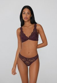 Burgunderfarbene Spitzen-Bralette mit breiten, gewellten Trägern und einem zentralen dekorativen Knopf, kombiniert mit einem passenden Spitzen-String, der ein ähnliches Muster aufweist.