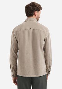 Man wearing a beige long-sleeve shirt facing away, with dark green pants, against a plain white background.