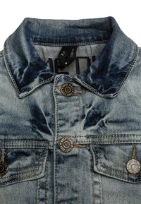 Denimjacka i en bleknad blå färg, med spetsig krage och metallknappar. Har två framfickor och en texturerad yta.