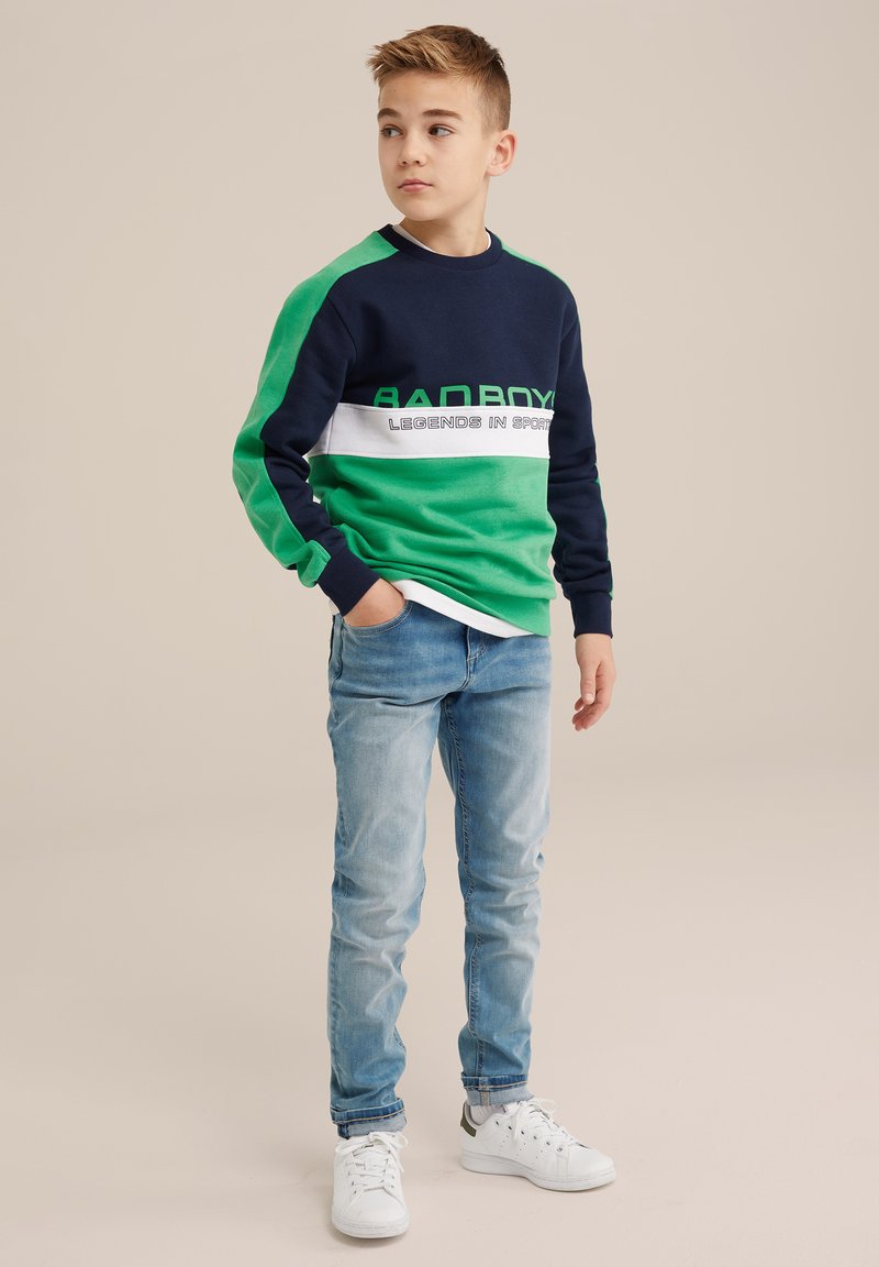 Pulover colorat în blocuri de navy și verde, cu textul "BAD BOY"; asortat cu blugi albaștri deschiși și adidași albi, având un material texturat.