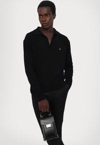 Black knit pullover with a collar, featuring ribbed shoulder details and a small logo. Paired with fitted black trousers and a sleek black handbag.