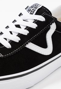 Svart semsket sneaker med hvite lisser, med et distinktivt hvitt bølgedesign på siden, rund tå og teksturert hvit gummisåle.