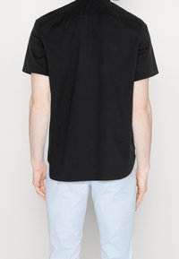 Man in een zwart shirt met korte mouwen en lichtblauwe broek, gezien van achteren tegen een effen achtergrond.