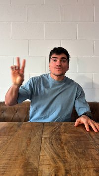 Muškarac u plavoj košulji sjedi za drvenim stolom, lijevom rukom drži tri prsta podignuta, dok je desna ruka naslonjena na stol.