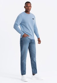 Sudadera azul claro con cuello redondo y detalle de etiqueta negra, combinada con jeans azul claro y zapatillas blancas.