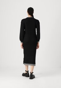 Crna haljina dugih rukava s uskim krojem, s rebrastim detaljima na izrezu i rubovima, te bočnim prorezima. Uparena s robusnim crnim cipelama.