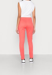 Kvinna klädd i korallrosa mjukisbyxor med vita sidostråkar, vit ärmlös topp och vita sneakers, står vänd bort.