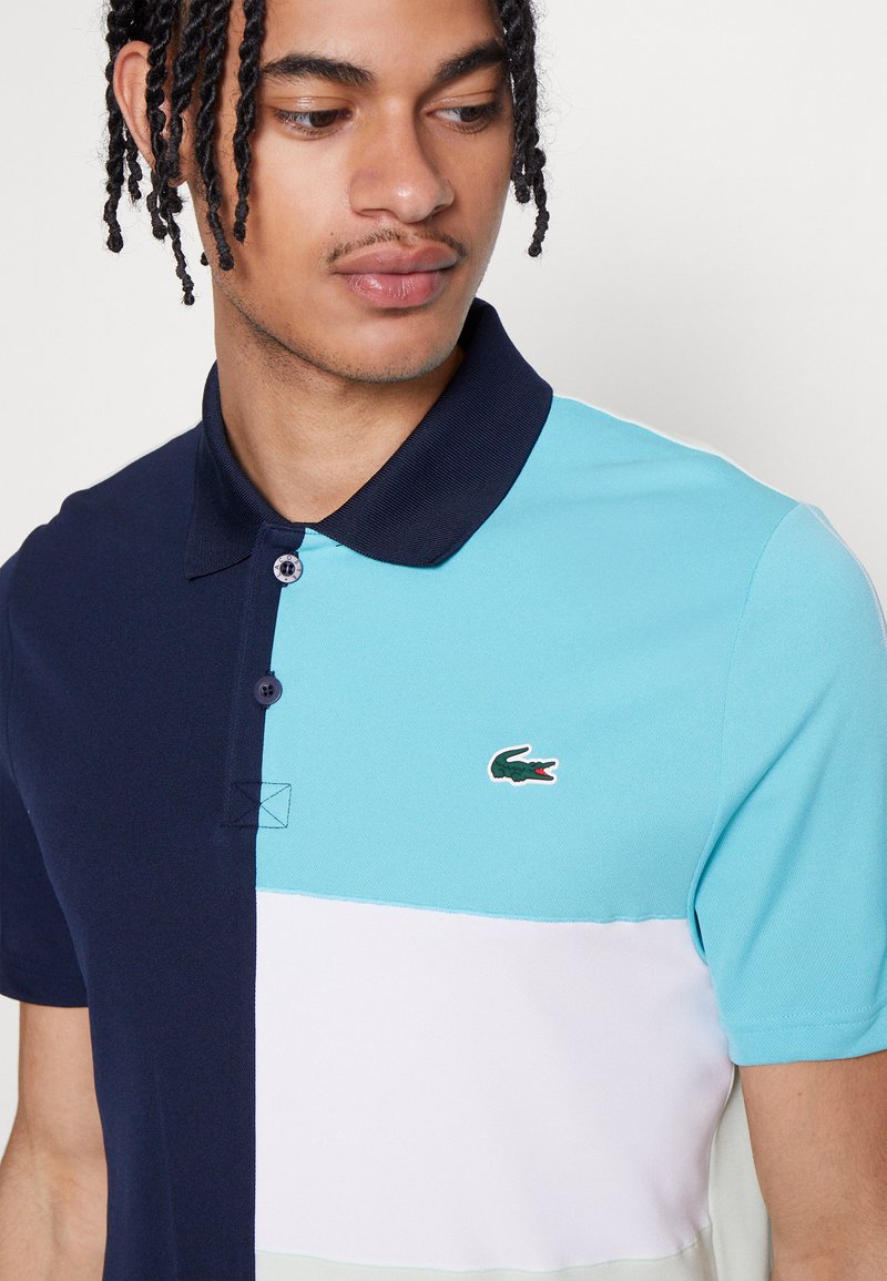 Homme avec des cheveux tressés portant un polo à manches courtes en navy, bleu clair et blanc avec un logo de crocodile vert brodé.