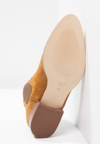 Brązowe zamszowe buty na obcasie z teksturowaną brązową podeszwą, oznaczone „MADE IN SPAIN” oraz rozmiarem „38”, o zakrzywionym kształcie i gładkich akcentach.
