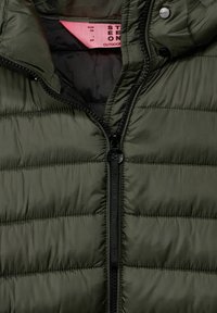 Mörkgrön puffjacka med quiltat mönster, utrustad med en svart dragkedja och tryckknappar. Insidan är fodrad med svart tyg och har en rosa etikett.