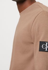 Beige Langarm-Sweatshirt mit einem strukturierten Waffelmuster. Verfügt über ein schwarzes Marken-Patch am Ärmel. Einfacher Rundhalsausschnitt.