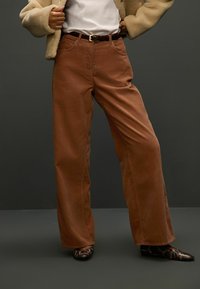 Braune Cord-Hosen mit weitem Bein und strukturiertem Finish, kombiniert mit einem schwarzen Gürtel. Das Outfit wird durch gemusterte Schuhe abgerundet.