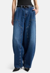 Wide-leg blue denim jeans featuring prominent stitching and metal button details, with a black belt and a textured surface.
