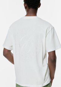 Maglietta bianca a maniche corte con un design del logo testurizzato sul retro, impreziosita da elementi decorativi come foglie di alloro e un banner curvo.