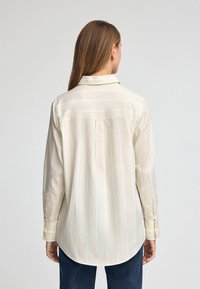 Camicia button-up gialla chiara con righe verticali bianche, maniche lunghe, colletto e orlo curvo. La texture sembra liscia e leggermente croccante.