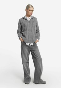 Szara bluza z kapturem na zamek, wykonana z dzianiny typu cable-knit, w połączeniu z luźnymi szarymi spodniami. Stylizacja zawiera warstwową białą koszulę z kołnierzykiem pod spodem.