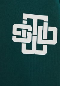 Una tela de color verde oscuro con una textura suave presenta un diseño de logotipo geométrico blanco prominente en el centro, creando un fuerte contraste visual.
