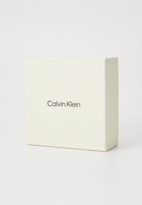 Boîte couleur crème de Calvin Klein avec une finition lisse, arborant un branding minimaliste dans une police moderne sur le devant. Design de forme carrée.