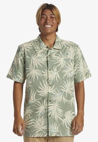 Camisa de manga corta abotonada en tejido verde con un estampado de hojas de palmera blancas y detalles en azul; cuello clásico y botones en la parte delantera.