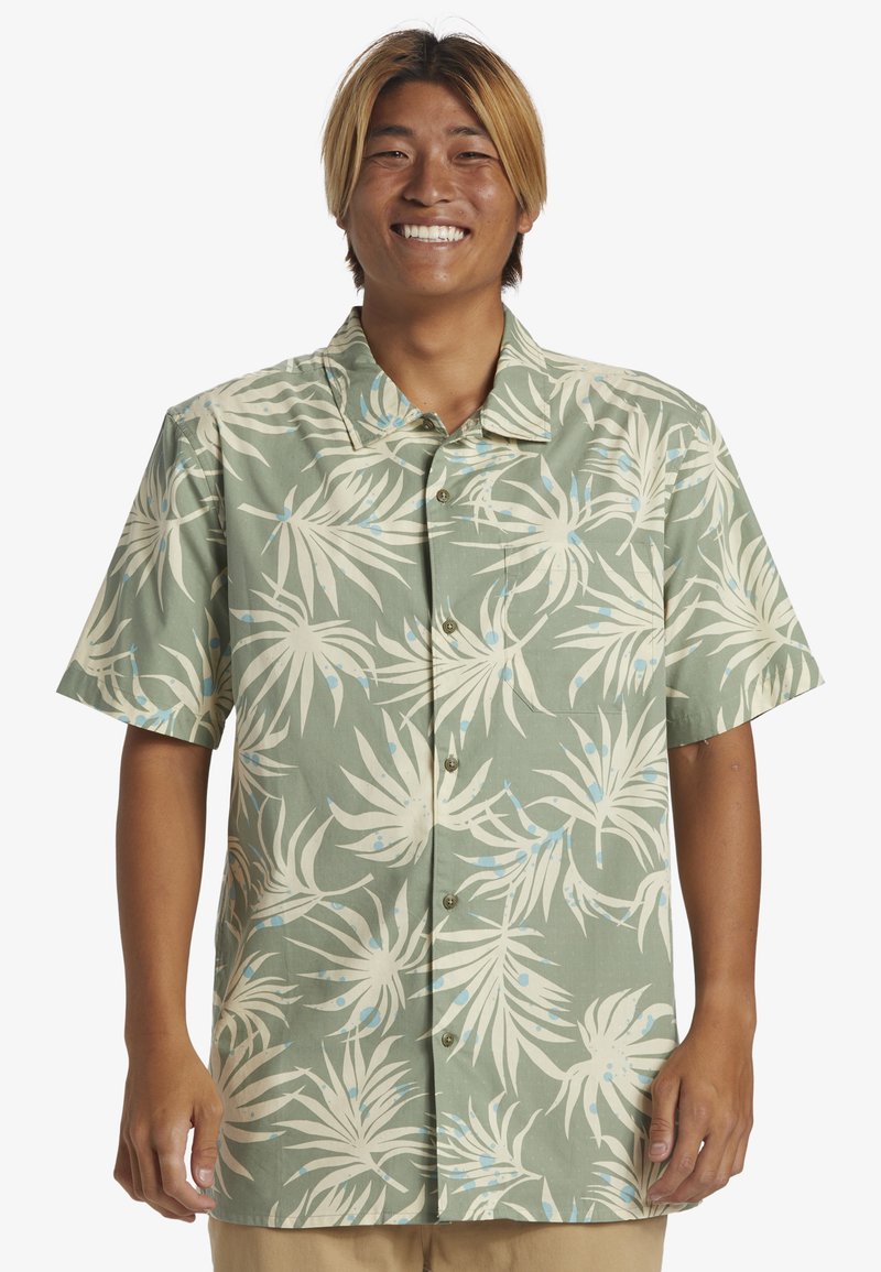 Camisa de manga corta abotonada en tejido verde con un estampado de hojas de palmera blancas y detalles en azul; cuello clásico y botones en la parte delantera.