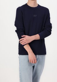 Marinblå långärmad skjorta med ribbad textur och rund halsringning, prydd med en diskret GAP-logotyp i mitten. Bärs med ljusblå jeans.