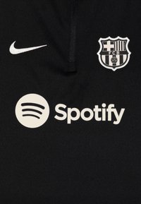 Fekete póló, fehér Nike logóval, FC Barcelona címerrel és jellegzetesen megjelenő fehér Spotify logóval. Az anyaga simának tűnik.