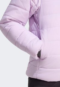 Ljuslila pufferjacka med detalj som visar en ärm och hand i fickan, vilket framhäver dess quiltade struktur och avslappnade stil.