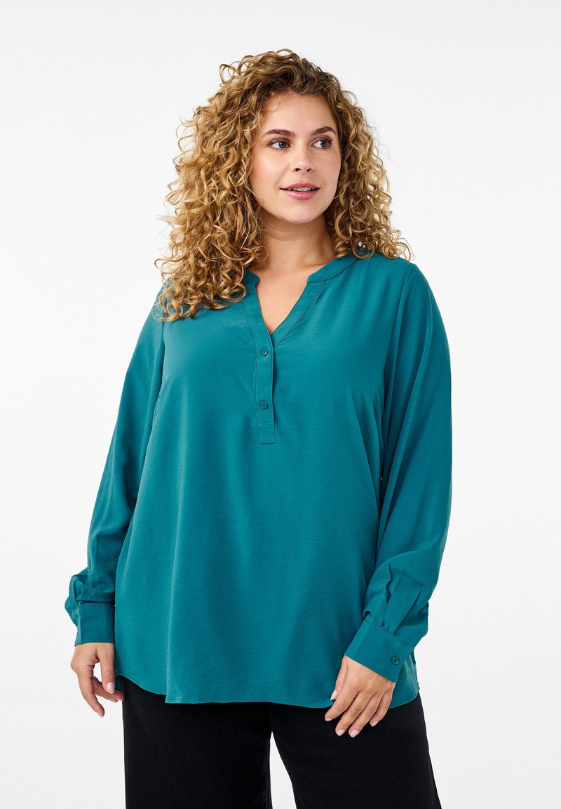 Tropfengrünes Langarmbluse mit V-Ausschnitt, drei-Knopfleiste, weicher Textur und leichter Wölbung am Saum. Kombiniert mit schwarzen Hosen.