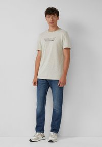 Hellbeige T-Shirt mit kurzen Ärmeln und bedrucktem Text, kombiniert mit blauen Jeans und grauen Sneakers, in einem einfachen, lässigen Design.