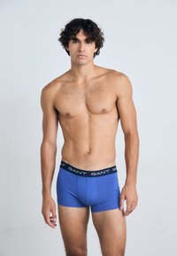 Boxer briefs de algodón azul con una cinturilla negra que presenta el logo de "GANT", exhibidos en un modelo sobre un fondo neutro.