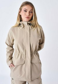 Beige vandtæt jakke med en tekstureret overflade, justerbar hætte, elastisk talje og to frontlommer; minimalistisk design uden synlig branding.