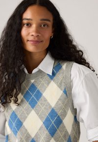 Gilet en laine gris avec un motif argyle bleu, blanc et jaune, porté par-dessus une chemise à col blanc. Texture douce, design sans manches.