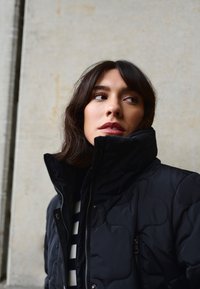 Čierna prešívaná páperová bunda s vysokým golierom, zipsom a bočnými vreckami, nosená cez pruhované tričko s viditeľnou textúrou a detailmi šitia.