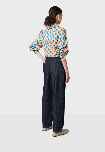 Camicia a bottoni con motivo, sfondo giallo e pois blu e rossi. Abbinata a pantaloni a gamba larga a righe scure e scarpe beige senza lacci.