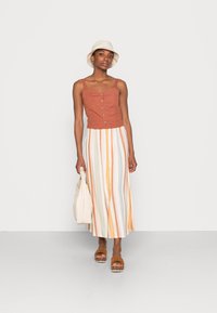 Top cropped de linho castanho com detalhes em botão, combinado com uma saia longa de riscas multicoloridas. Chapéu de balde bege e sandálias de plataforma castanho claro.