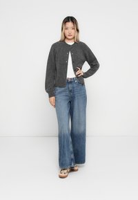 Grauer Wollmischung-Pullover mit rundem Halsausschnitt, vorderen Knöpfen und langen Ärmeln, kombiniert mit weit geschnittenen blauen Jeans und gemusterten Schuhen.