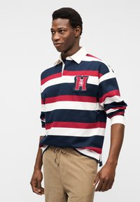 Άνδρας που φοράει μακρυμάνικο πουκάμισο ράγκμπι με κόκκινες, λευκές και ναυτικές ρίγες και μπεζ παντελόνι, στέκεται με τα χέρια χαλαρά στα πλευρά του.