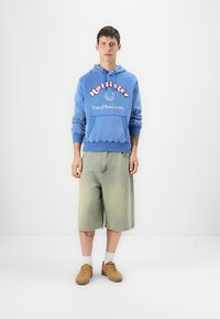Blue cotton hoodie with graphic text, paired with light green denim shorts. Model wears brown sneakers and white socks. Plain background.