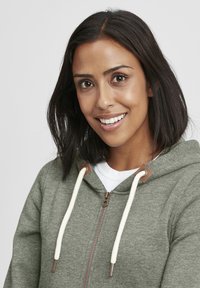Grønn zip-up hettegenser med kremfargede snorer, brune detaljer og foret hette, iført over en hvit skjorte. Stoffet ser mykt og strukturert ut.