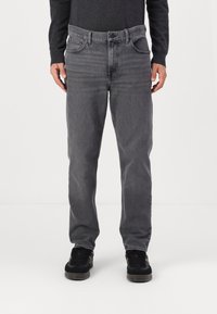 Hombre vistiendo jeans rectos grises, suéter negro y zapatillas negras, de pie contra un fondo blanco liso.