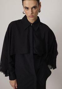 Black layered blouse with a structured collar, featuring sheer ruffled sleeves and a subtle gold button at the collar. Smooth fabric texture.